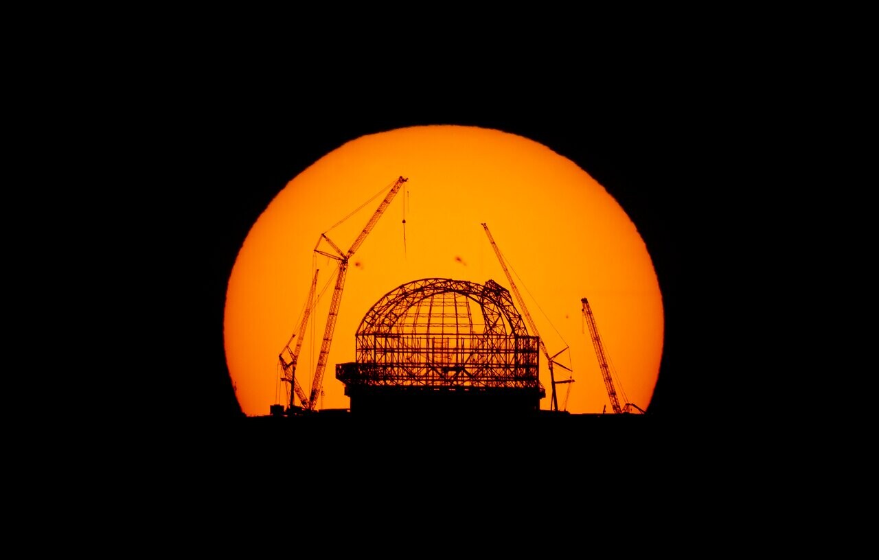 The huge orange disc of the Sun dominates the picture against a black background, due to the way the image was taken. The Sun has nearly fully risen above the ground and is just cut off from being a perfect circle at the bottom. Much like shadow puppets on a screen, the dark outline of the criss-crossed steel structure of the ELT dome is positioned at the centre of the Sun. There are large cranes leaning in over the central dome, also only distinguishable as dark outlines.