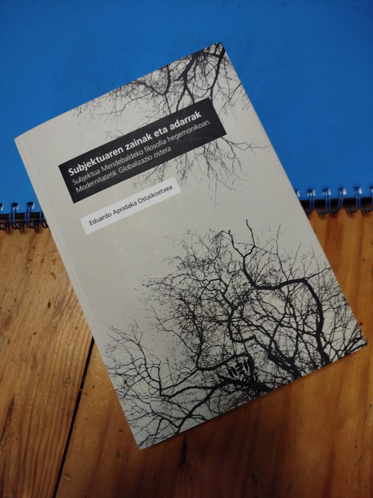 Eduardo Apodaka Ostaikoetxea filosofoaren liburua ikusten da egurrezko mahai batean, erdizka koaderno urdin baten gainean. Liburua grisa da eta adar-sustrai formak ikusten dira beltzez, liburuaren titulua da: Subjektuaren zainak eta adarrak. Subjektua Mendebaldeko filosofia hegemonikoak Modernitateak Globalizazio ostera.
