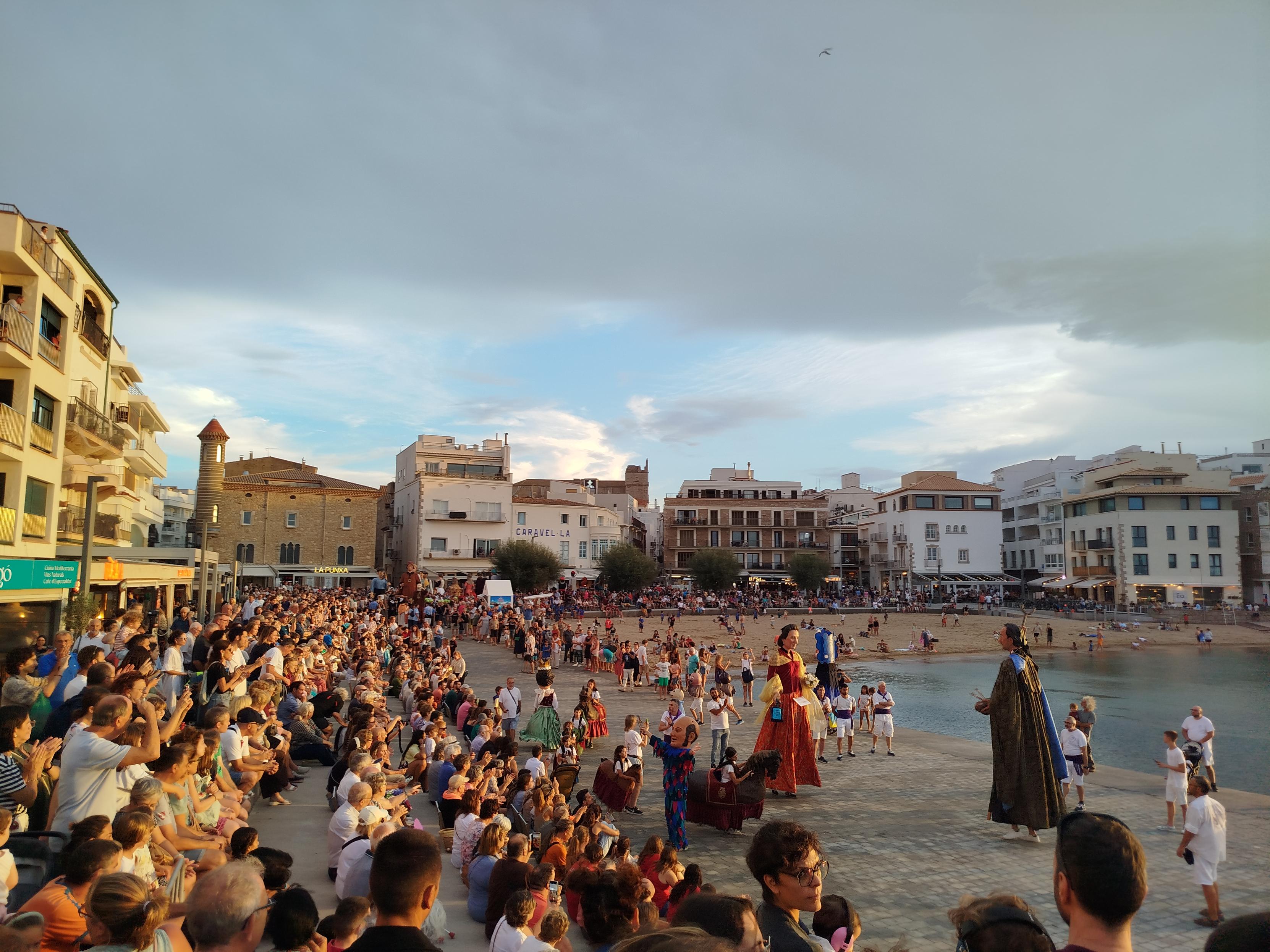 Bi erraldoi dantzan, konpartsa kideak inguruan eta jendea eguzkitan ikusten L'Escalako Riban, portua eta hondartza dituena. Bi erraldoi dantzan, konpartsa kideak inguruan eta jendea eguzkitan ikusten L'Escalako Riban, portua eta hondartza dituena.